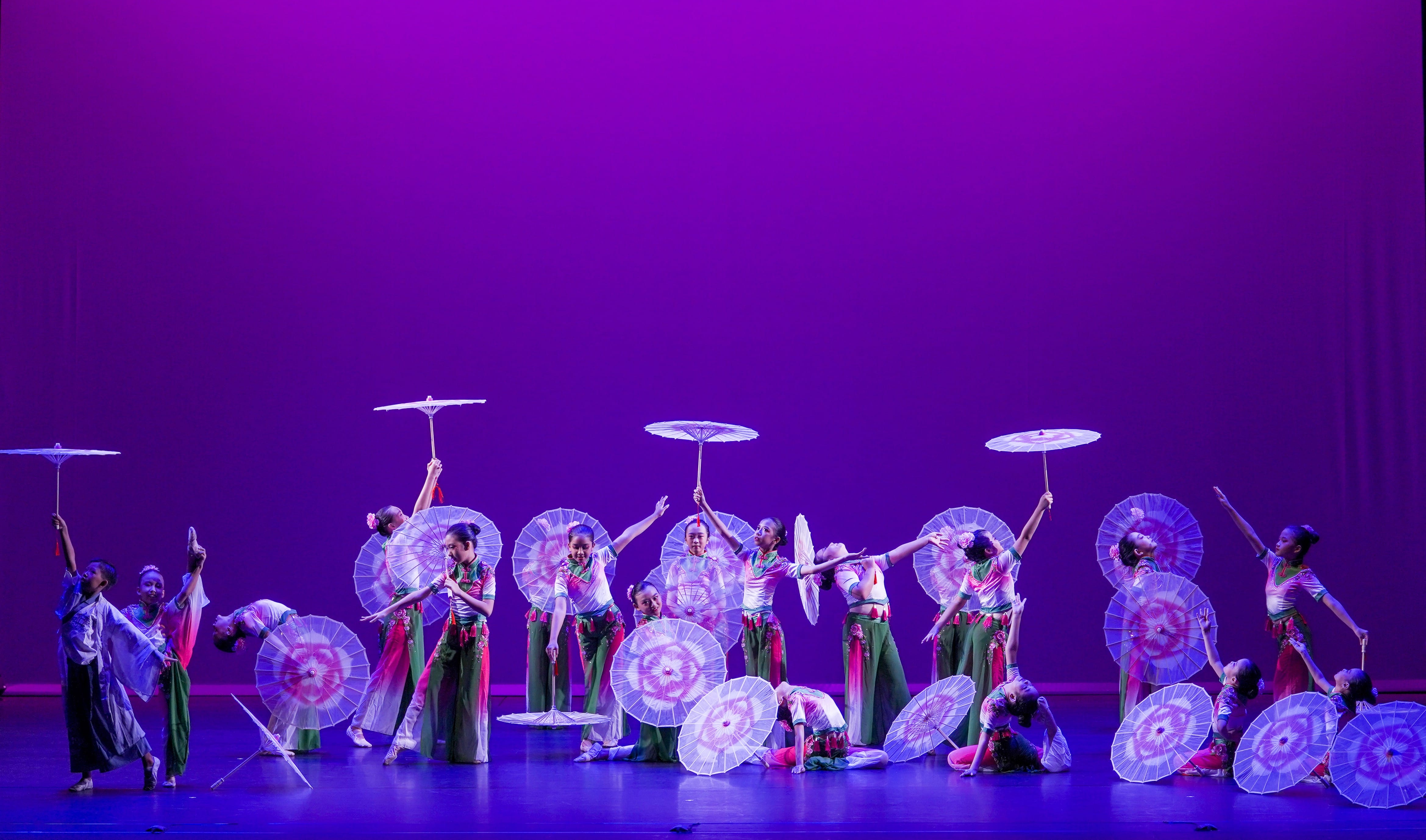 Dancers in traditional Chinese costumes perform with decorative parasols on a purple-lit stage.